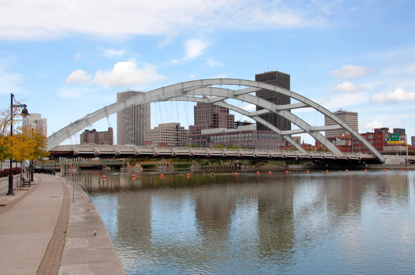 A wide view of a white tied-arch bridge crossing a calm river toward the Rochester city skyline, viewed from a paved waterfront walkway.
